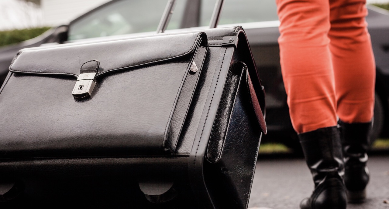 Une personne portant des pantalons rouges et des bottes noires se dirige vers une voiture avec une valise à roulettes noire