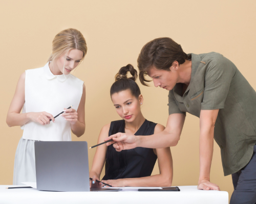 Trois personnes se tiennent devant un ordinateur portable. Une femme est debout, une femme est assise et un homme est debout pointant l'écran de l'ordinateur portable avec un crayon.