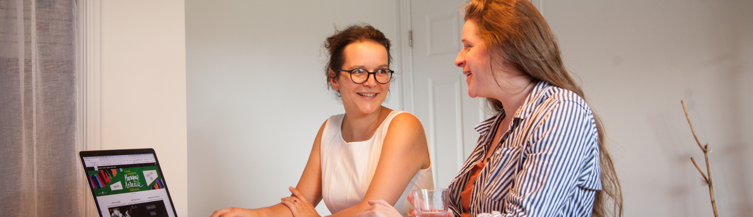 Deux femmes se regardent dans les yeux en souriant. Devant elles, un ordinateur est ouvert sur un site web 