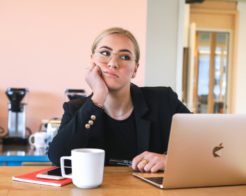 Une femme, d'un air ennuyé, tient sa tête avec sa main sous le menton. Sur la table, il y a un cahier, une tasse et un ordinateur portable.